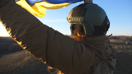 Female soldier of ukrainian army holding waving flag of Ukraine. Girl in military uniform and helmet lifted up yellow-blue flag. Victory against russian aggression. Invasion resistance concept. Slowmoの写真素材