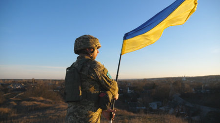 Soldier of ukrainian army stands at hill and holds waving flag of Ukraine. Man in military uniform...
