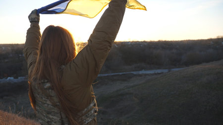 Female ukrainian army soldier holding waving flag of Ukraine. Girl in military uniform lifted up yellow-blue flag against blue sky. Victory against russian aggression and the end of the war. Close upの写真素材