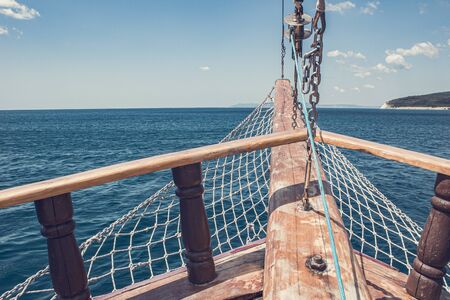 View of the island from the old wooden ship. The bow of a vintage ship. The wooden stem of a ship, the dangling net, the furled sails and wooden beams of an ancient ship sailing past the land.の写真素材