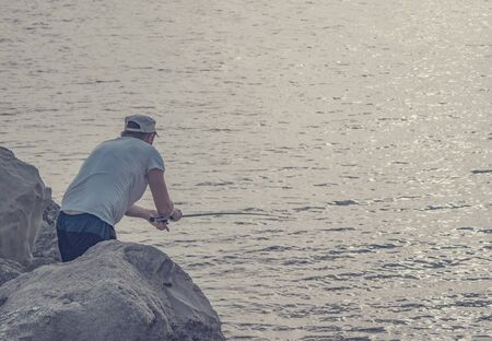 A grown man throws a fishing rod into the sea. Fisherman fishing from rocks and stones in the oceanの写真素材