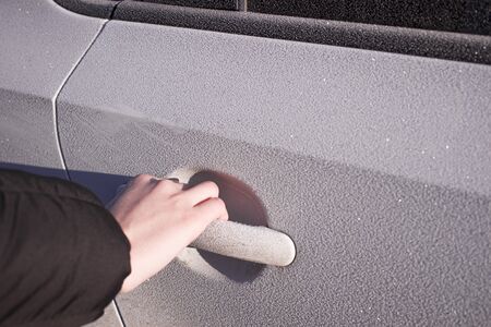 A person holds a frozen car handle in winter. A man opens the door in a white car covered in hoarfrost morning frosts.の写真素材