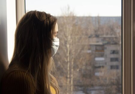 A girl in a mask sits on the windowsill and looks out the window at the city. A woman in a medical mask sits in quarantine. The concept of quarantine and self-isolation.の写真素材