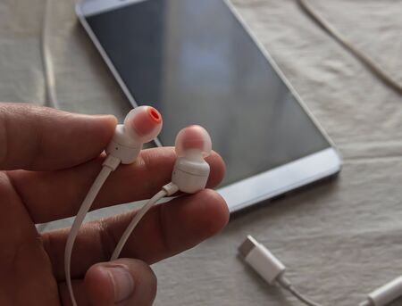 A man is holding a phone earphone in his hands. Headphones in hand on the background of a smartphone. The phone is on the table.の写真素材