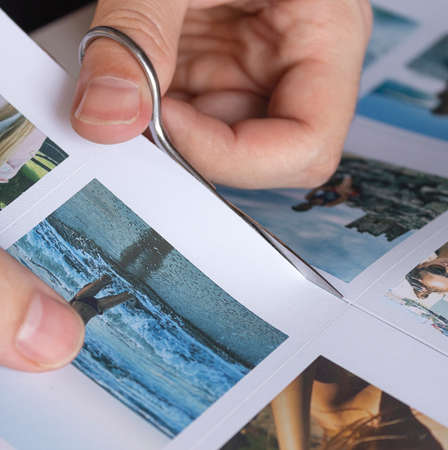 A man cuts out photos with people in close-up. A man uses scissors to cut printed photos for an album. A person holds a photo in his hands against the background of other photos.の写真素材