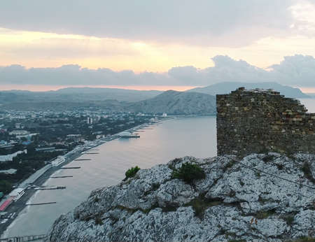 A ruined brick building on top of a cliff with a view of the resort town in cloudy weather and a pale pink skyの写真素材