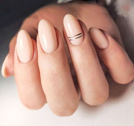 Gentle gel polish of beige color on almond-shaped nails with silver stripes. Nude manicure with shiny stripes design. Pastel color coating on the nails.の写真素材