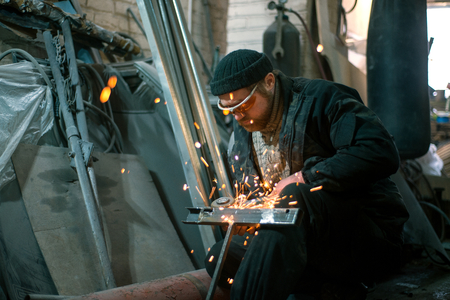 The worker cuts the steel with an electric grinding wheel on his kneeの写真素材