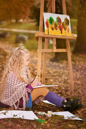 little girl artist paints a picture on an easel in autumn in the parkの写真素材