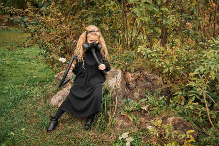 little girl in respirator mask and backpack with plants behind her back holds white rose in her hands. the child is sitting on tree stump ecology concept clean air and apocalypseの写真素材