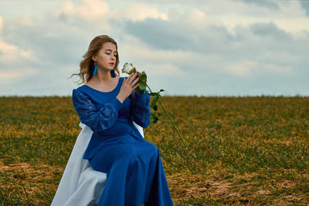 romantic woman portrait. female in blue dress holds white rose in her hands while sitting on chair in the fieldの写真素材