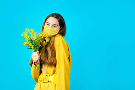 portrait of a teenage girl in a glamorous mask on her face with mimosa flowers in her hands. blue...