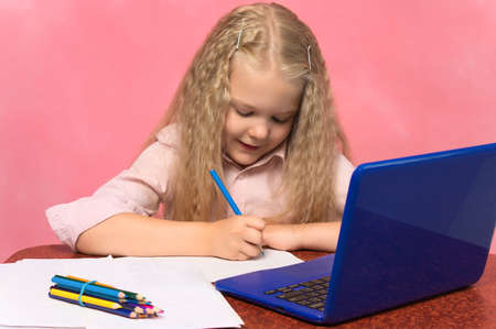little schoolgirl with pencils writes at a table with a laptop. pink background