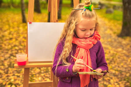 little girl artist with a brush and paints in her hands draws in autumn in the parkの写真素材