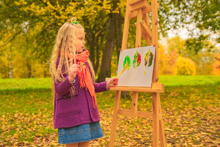 little girl artist with a brush and paints in her hands in autumn in the park draws a landscape with leaves on canvasの写真素材