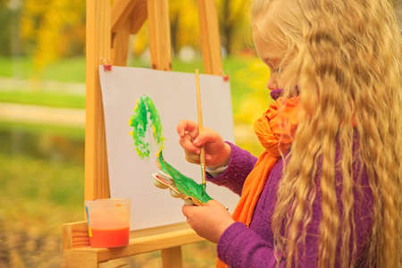 little girl artist with a brush and paints in her hands in autumn in the park draws a landscape with leaves on canvasの写真素材