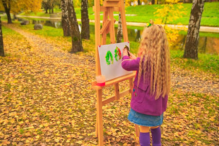 little girl artist with a brush and paints in her hands in autumn in the park draws a landscape with leaves on canvasの写真素材