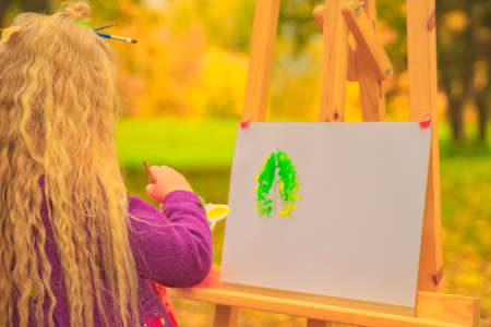 little girl artist with a brush and paints in her hands in autumn in the park draws a landscape with leaves on canvasの写真素材