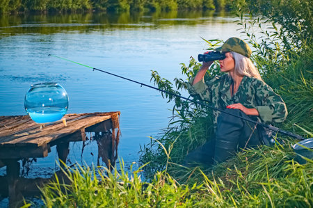 Woman fisherman carefully looks through binoculars on the river bank. Summer, hobby, vacationの写真素材