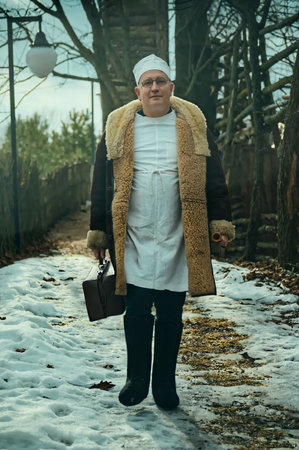 Male doctor veterinarian with a suitcase walks through a ranch village in winter in warm clothes and felt bootsの写真素材