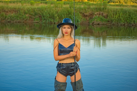 Attractive woman in a leather swimsuit wearing a hat with a fishing rod in her hands. Lady fisherman fishing on the river bankの写真素材