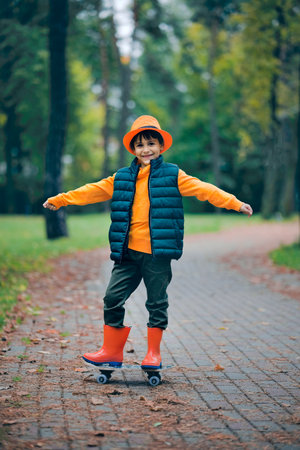 Fun portrait of a boy on a skateboard in the park in the fall. Kid in a hat and orange rubber boots.