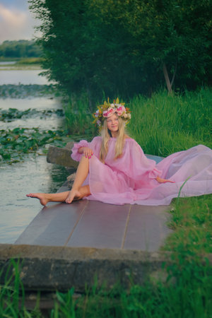 A young teenage girl sits on a pier by the riverside wearing a pink dress and a wreath on her head. Summer. concept inspirationの写真素材