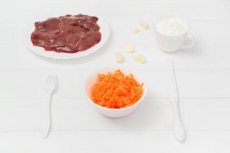 Fresh raw chicken liver and grated carrots in a plate and garlic cloves, Sour cream gravy boat. White wood background. light keyの写真素材