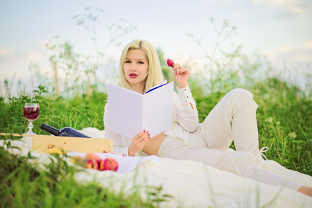 A beautiful blonde woman is reading a book and eating strawberries. Lady relaxing on a picnic, lying on a plaid on the green grassの写真素材