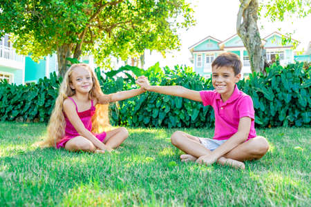A little smiling girl and a boy sitting on the grass in a summer garden and holding hand, looking straight in the camera, the concept of friendshipの写真素材