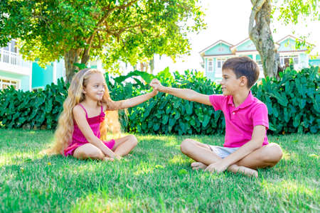 A little smiling girl and a boy sitting on the grass in a summer garden holding hands and looking at each other, concept of friendshipの写真素材