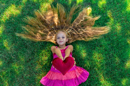 A cute little girl with long wavy hair lying on the grass, looking up and holding a red velvet heartの写真素材
