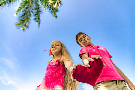 Little smiling girl and boy staying under a palm tree and holding red heart, copy spaceの写真素材