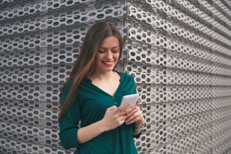 Young smiling female with white smart-phone in hands on metal panel background