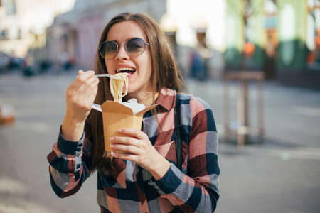 Beautiful smiling young woman in sun glasses eating fast food on cities background