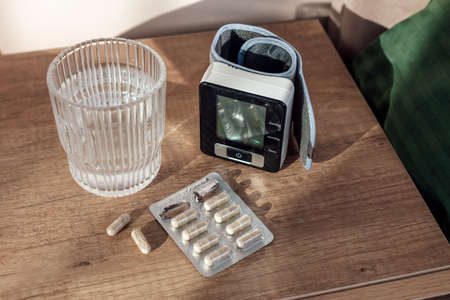 Pills capsules, glass of water and digital Blood Pressure Monitor on wooden background