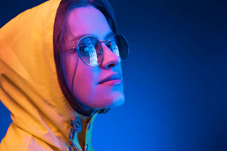 Neon close up portrait of young woman round sunglasses yellow raincoat on dark blue backgroundの写真素材