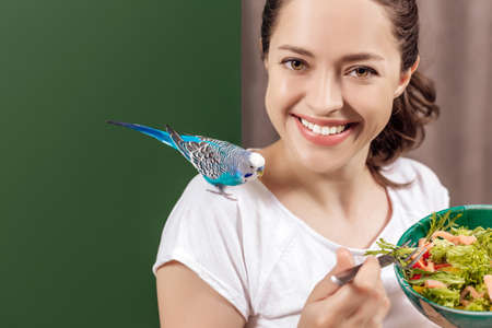 Portrait of a young cheerful woman with blue wavy parrot eating salad at the table full of healthy...