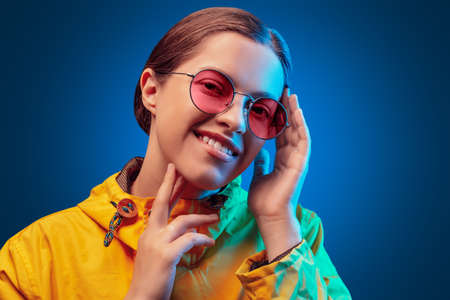 Portrait of fashion young girl in yellow raincoat and round sunglasses in neon light on blue...