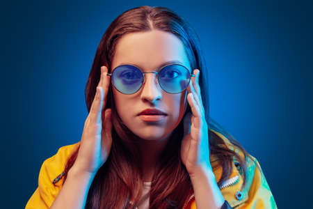 Portrait of fashion young girl in yellow raincoat and round sunglasses in neon light on blue backgoundの写真素材