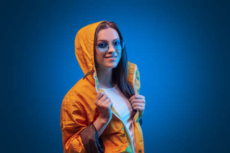 Portrait of fashion young girl in yellow raincoat and round sunglasses in neon light on blue backgoundの写真素材