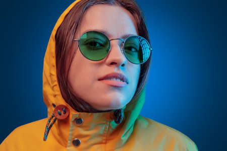 Portrait of fashion young girl in yellow raincoat and round sunglasses in neon light on blue...