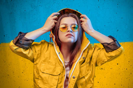 Young woman in yellow raincoat and round color glasses in ukrainian flag backgroundの写真素材