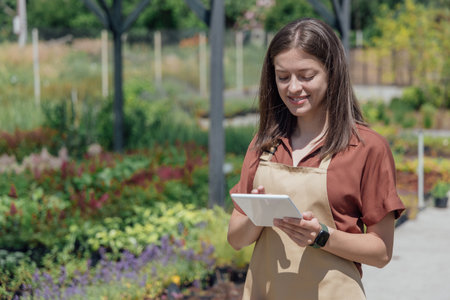 A young female gardener holds a gadget and checks information on plants in a garden center
