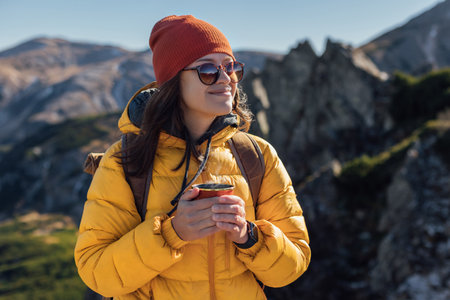 Portrait of a happy woman hiker in yellow down jacket standing with cup f tea on the slope of mountain ridge against mountainsの写真素材