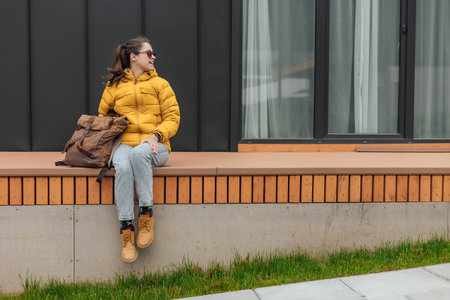 Young female in yellow down jacket with leather retro tourism backpack have a rest in nature background with cabin house behindの写真素材