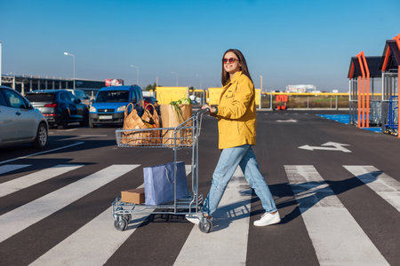 Woman in yellow jacket goes crosswalk from a shopping mall with shopping cart with a groceriesの写真素材
