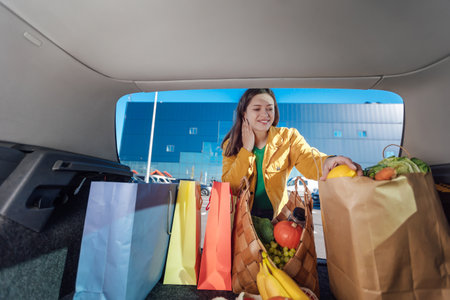 Woman in yellow jacket puts shopping bags with a groceries into a car trunk