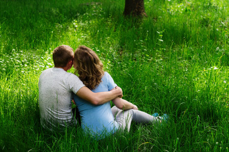 Rear view of couple of incognito girlfriend and boyfriend sitting in grass on ground embracing and kissing. Looking each other face to face.の写真素材