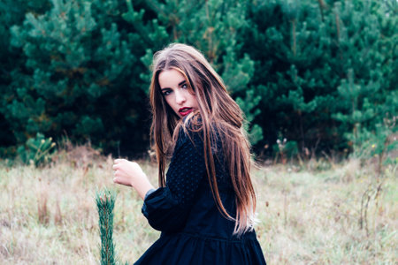 Portrait of turned back beautiful brunette girl with long hair and evening make up, posing and looking at camera over shoulder. Wearing black blouse. Pine forest at background.の写真素材